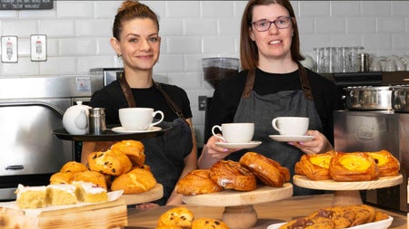 Two people standing behind a counter with pastries on display holding cups and a tray