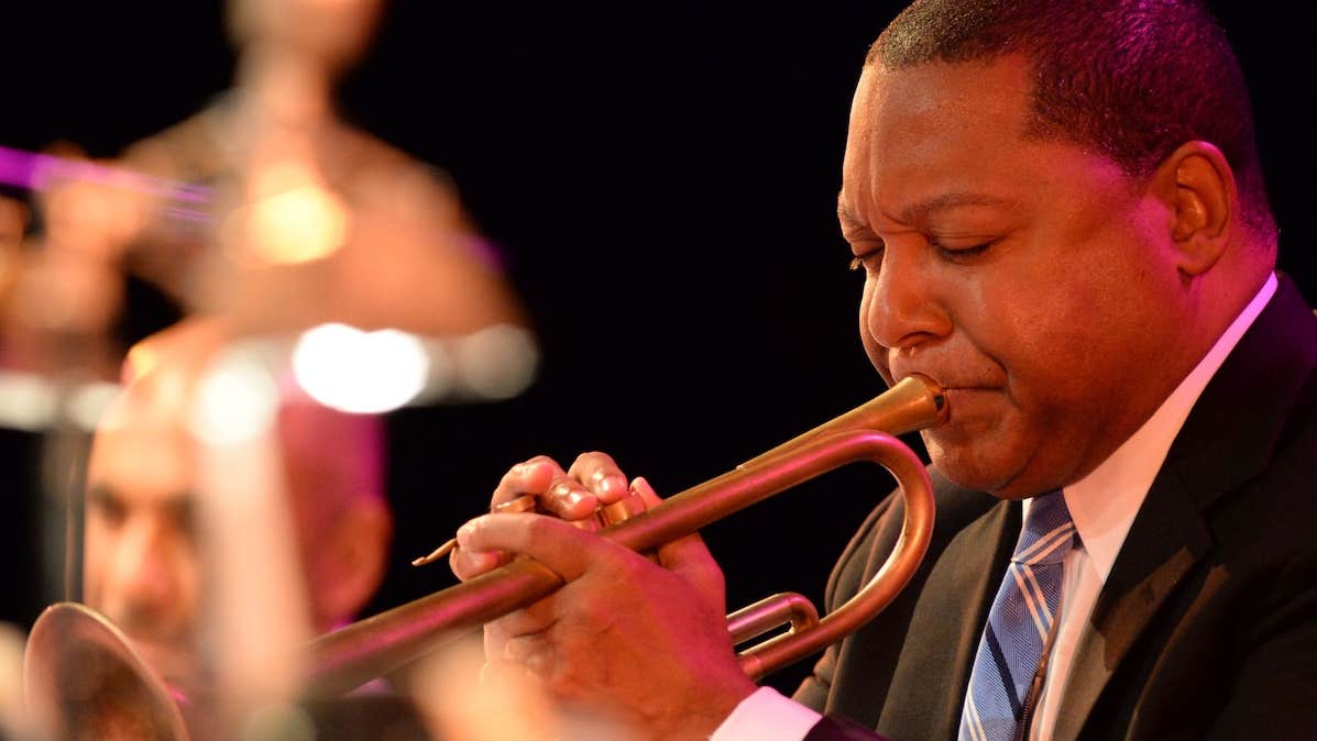 Close up view of man in suit and tie playing a trumpet with blurred orchestra in background.
