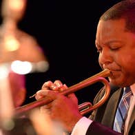 Close up view of man in suit and tie playing a trumpet with blurred orchestra in background.