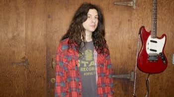 A person with long dark hair and checked shirt, standing beside a guitar hanging on the wall.