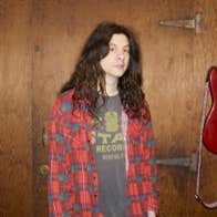 A person with long dark hair and checked shirt, standing beside a guitar hanging on the wall.
