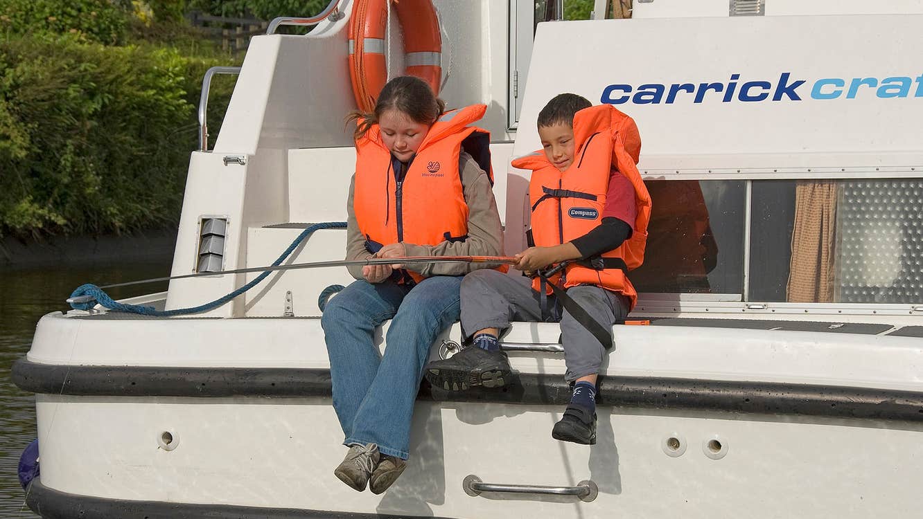Two kids wearing orange lifejackets sit on the back of a Carrickcraft cruiser