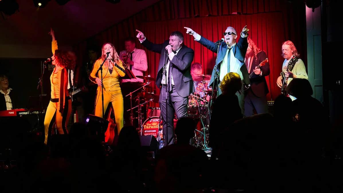 A group of 11 men and women singing and playing instruments with passion on a small stage.