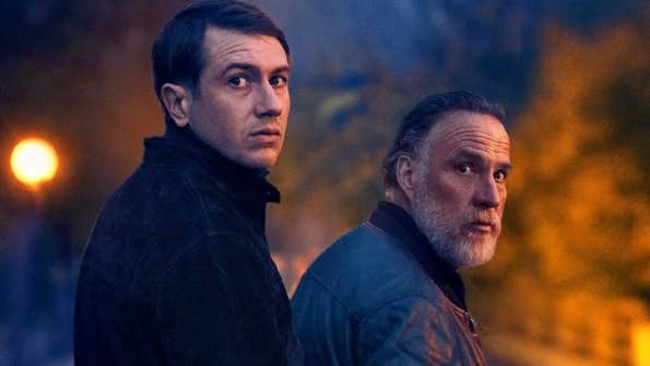 2 men in coats outside at night looking back over their right shoulders at the camera.