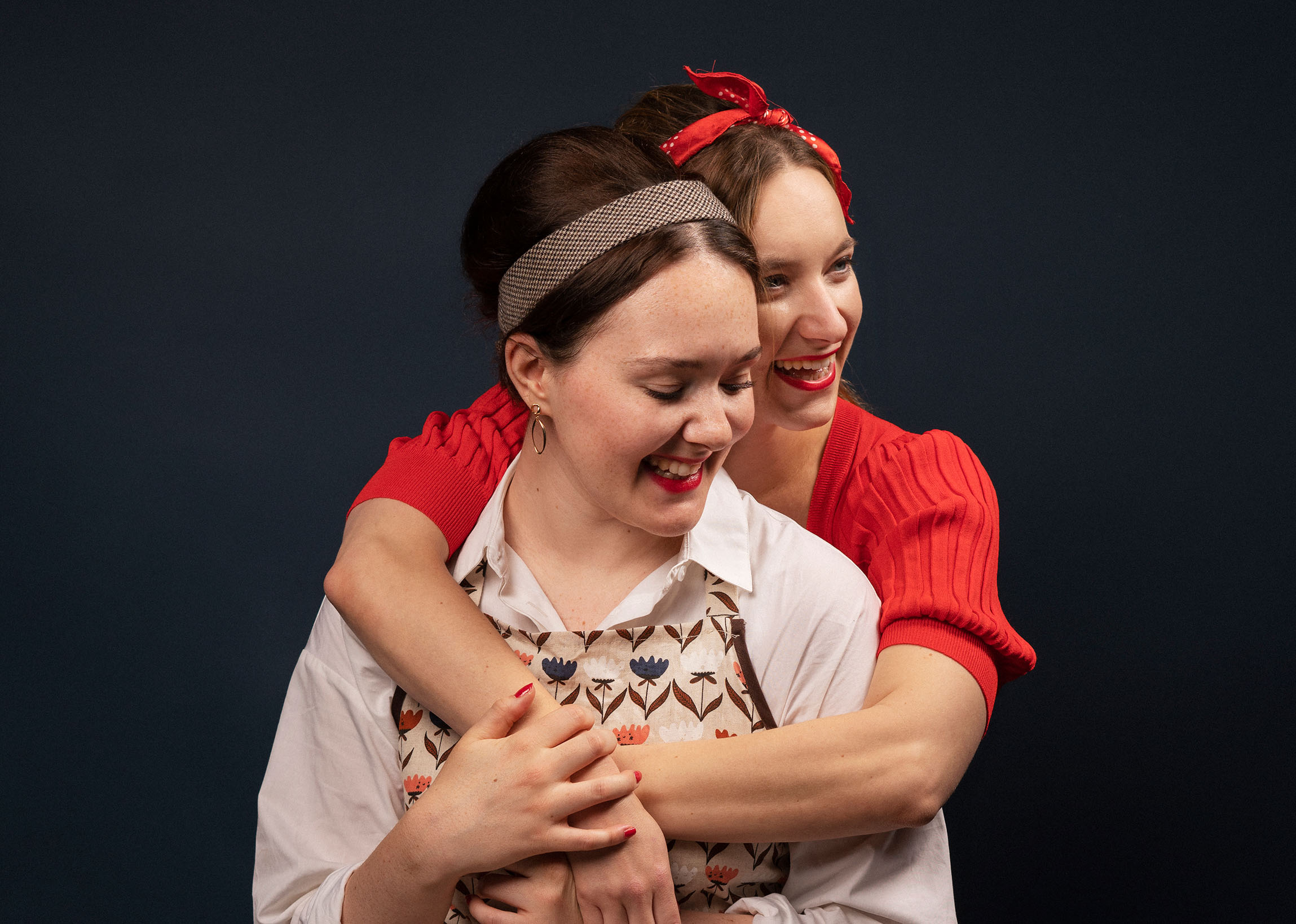 A photograph of two women in 1960s dresses hugging, one is wearing a red jumper, the other one a white shirt and an apron