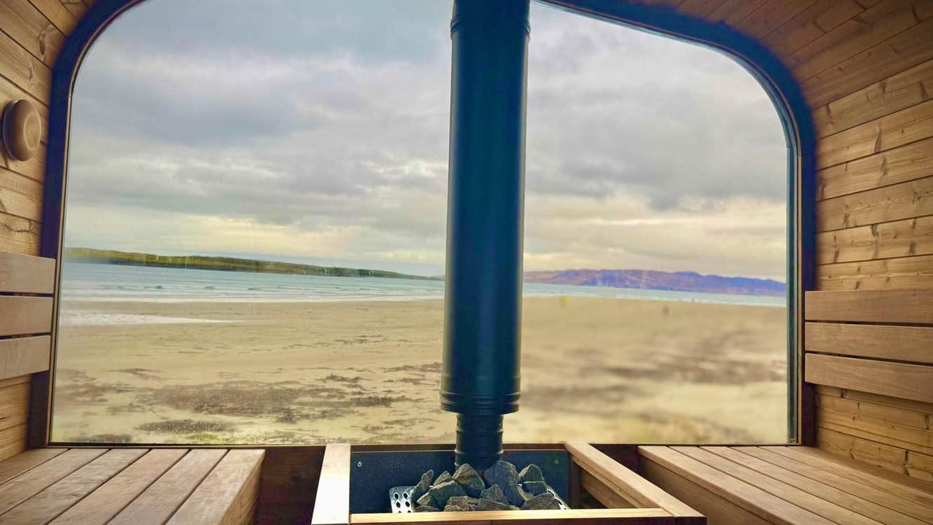 Interior of a sauna with wooden seating and a window with a view onto a sandy beach