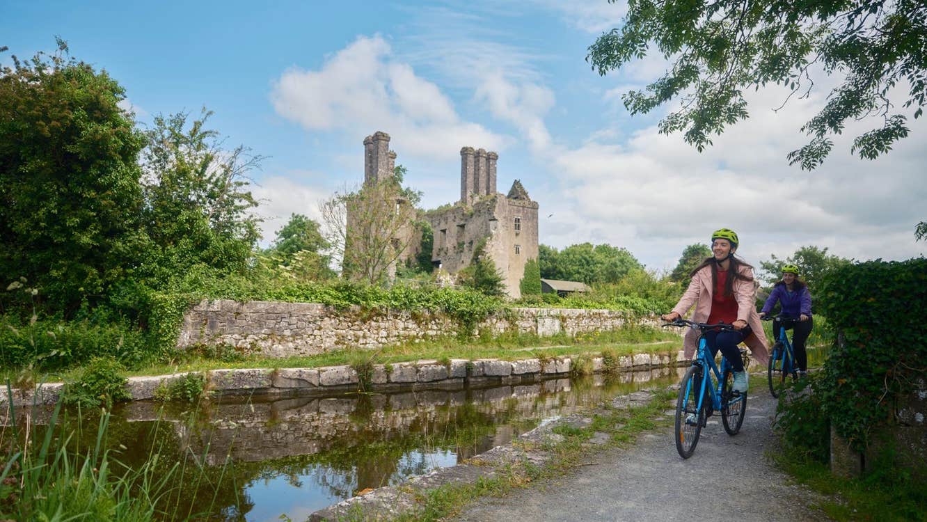 Two cyclists on a path by a river with the ruins of a castle in the background