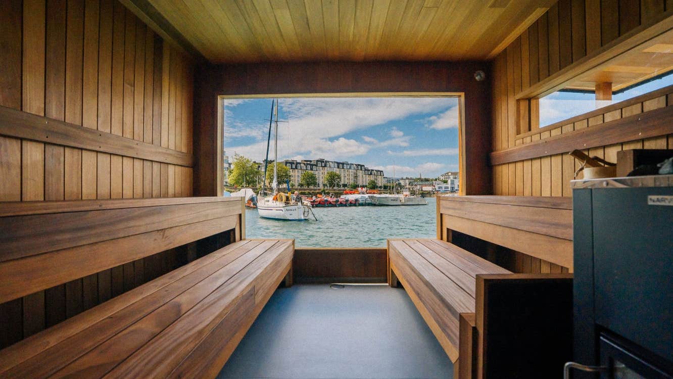 Wood lined sauna interior with bench seating overlooking a harbour through a large open window
