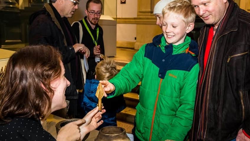 A adult and child touching the pointed end of an arrow head being help up by a woman seated at a table with other ancient artifacts.