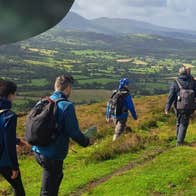 Rear view of people in hiking gear with rucksacks walking with distant view of hills and fields.