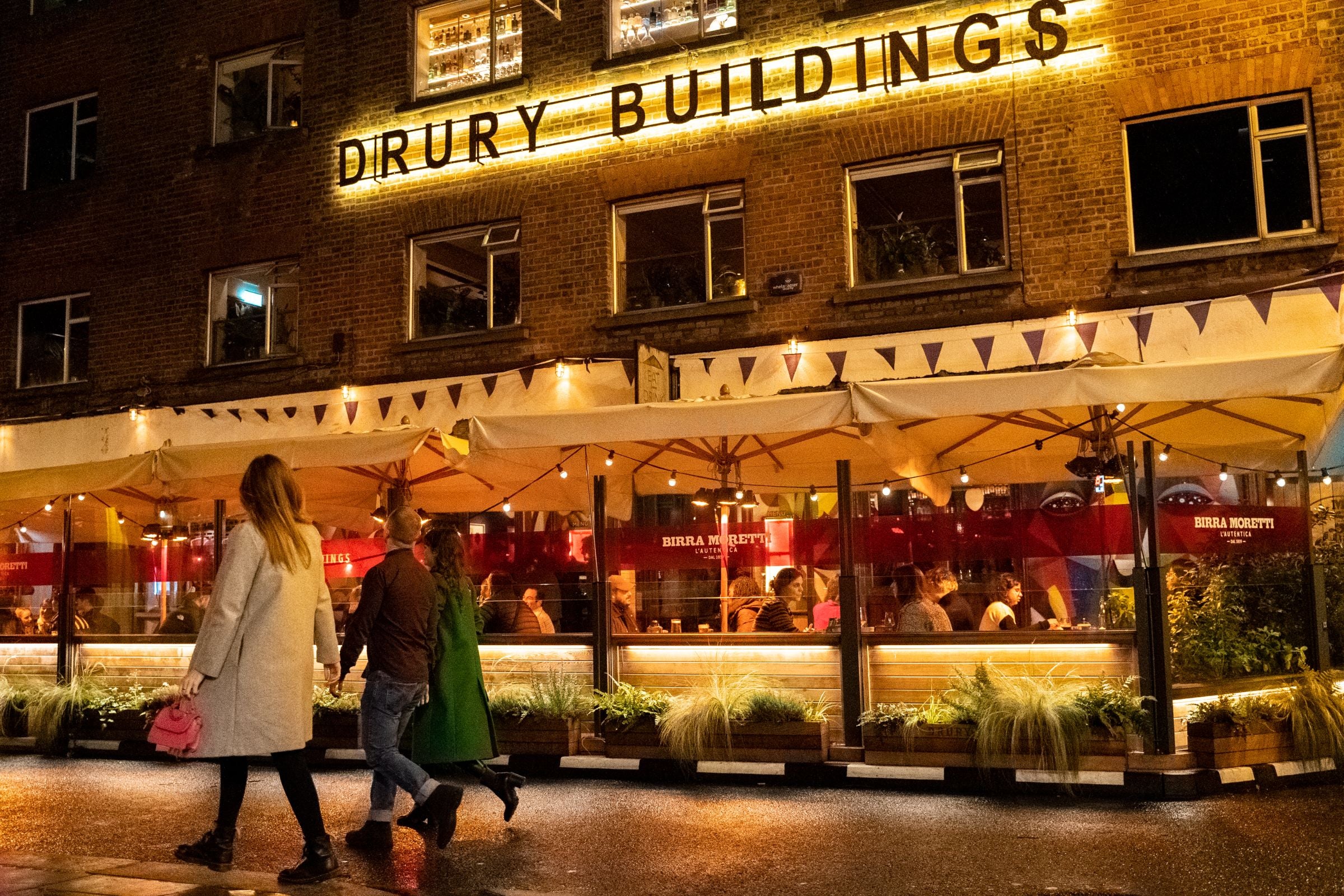 A group of friends walking by Drury Buildings on a night out with people sitting out on the heated patio.