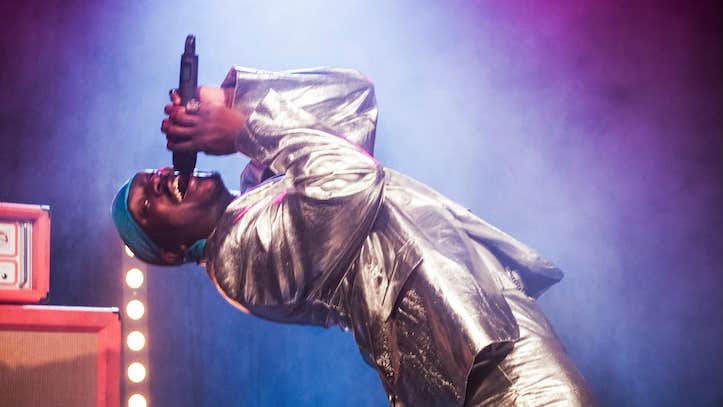 A man in shiny suit is singing into a hand held mic, bending backwards.