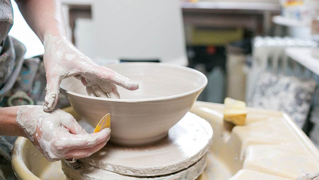 Clay covered hands working on a clay bowl on a potter's wheel.