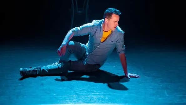 A man in denim is sat on the floor of a stage looking away to his left.