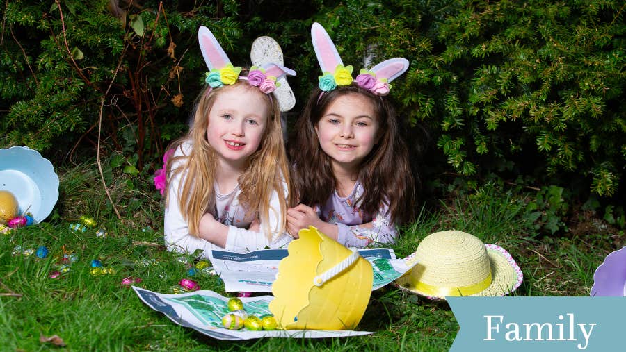 2 young children wearing white rabbits ears are laid down on grass with easter bucket and eggs in front of them and a yellow hat.
