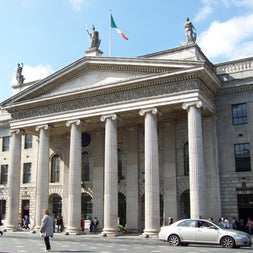 Cream coloured building with six columns and the Irish flag flying above on a street