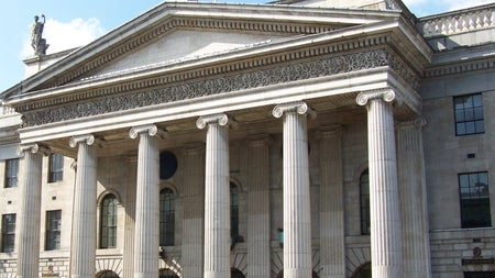 Cream coloured building with six columns and the Irish flag flying above on a street