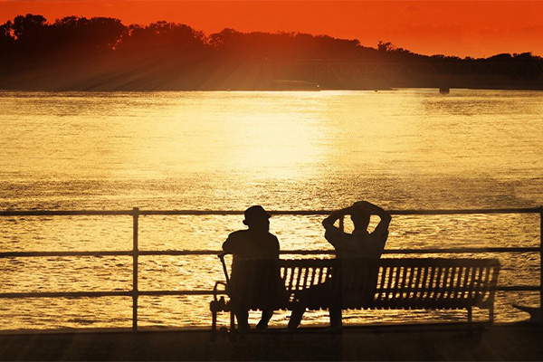 TUESDAYS WITH MORRIE BY MITCH ALBOM. Outlines of 2 figures sitting on a bench overlooking water with trees in distance, all at sunset with tones of black, yellow and red.
