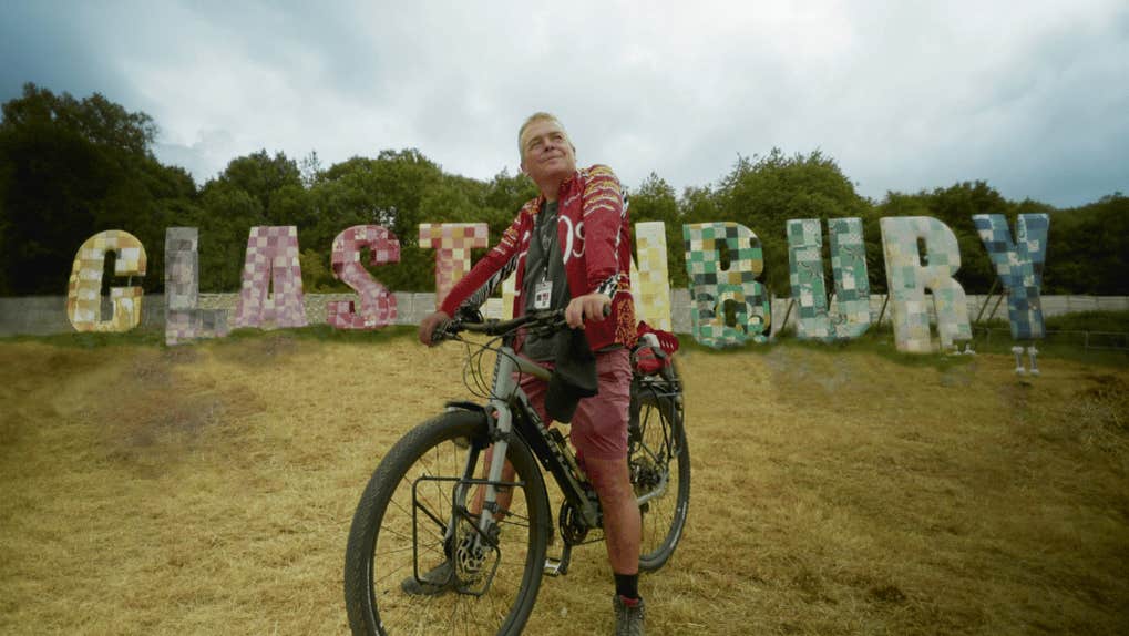 A man looking wistful on a bike with large letters in the background spelling out a name of a town