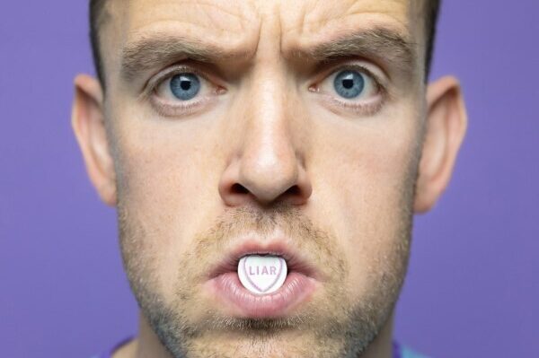 Face of a man looking concerned with a sweetheart sweet in between his lips with word liar on, against plain purple background.