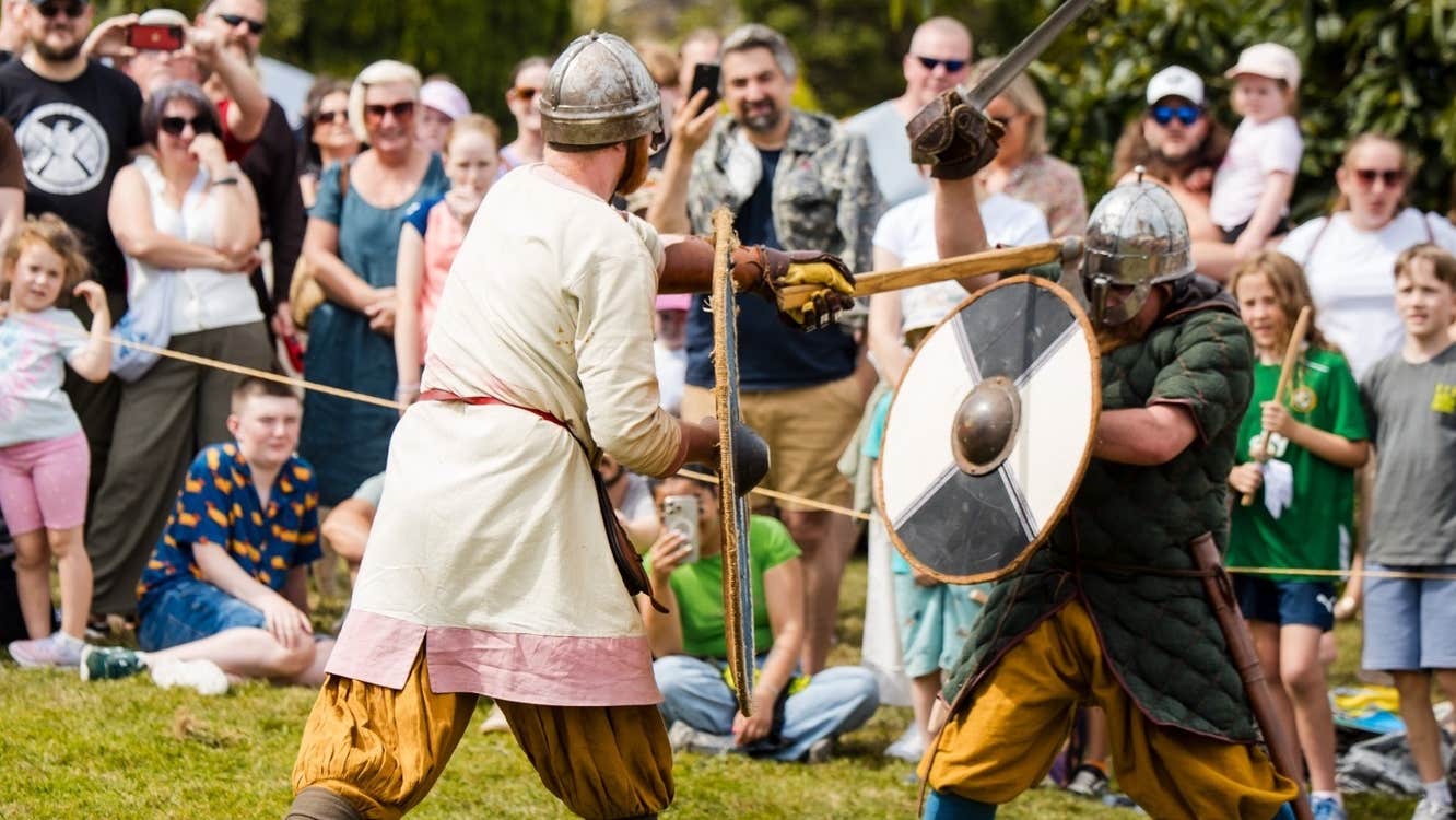Viking Battle Re-enactment, 2 men in viking style clothes with metal helmets fighting with round shields and wooden swords with crowd watching on.