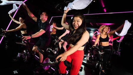 An spin class under neon lights with riders waving white towels