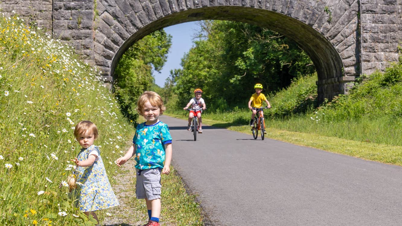 Two kids on bikes along the Old Rail Trail Greenway along with two kids that are picking flowers on the side of the trail