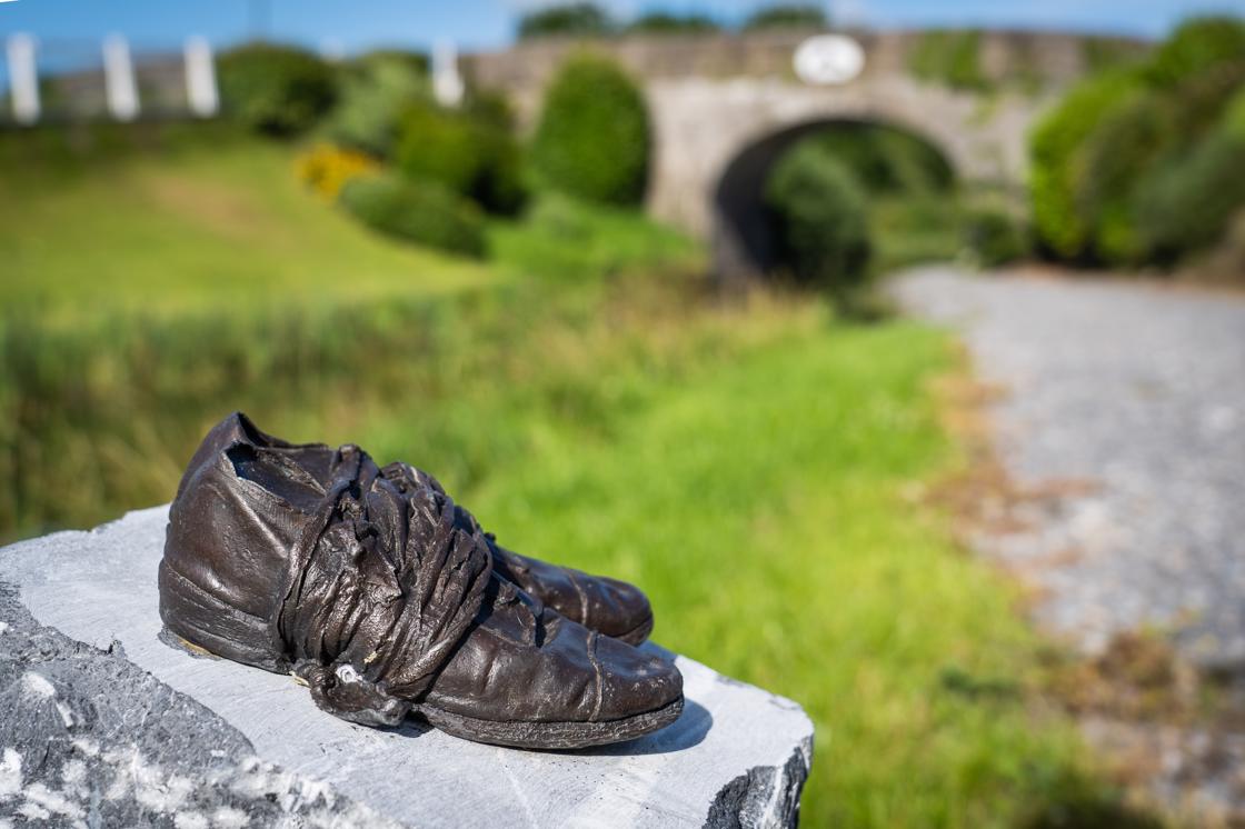 A bronze pair of old style shoes are on top of a grey stone plinth outdoors with a blurred large building in the background.