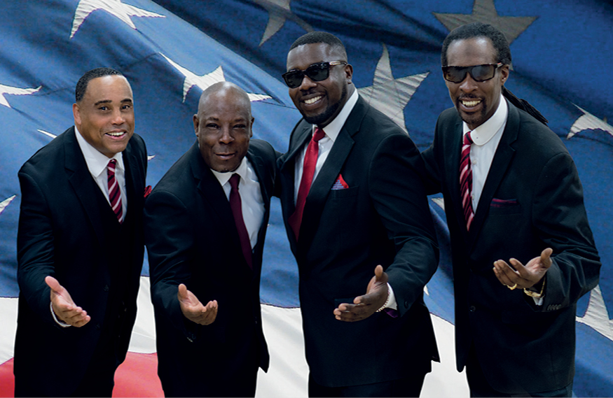 The Drifters Tribute Show. 4 smiling men in a line in suits, each with one hand outstretched.