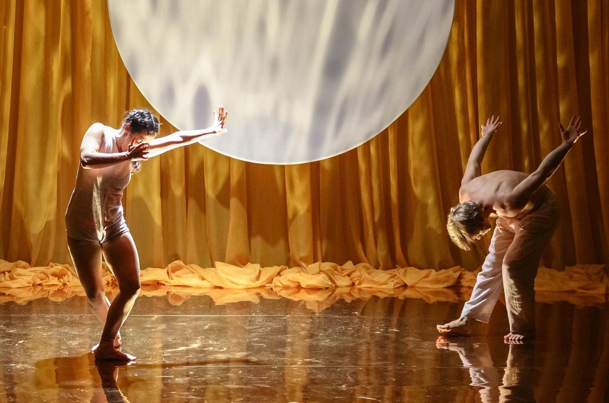 2 dancers on a stage with golden yellow curtains in the background with round white moon shape on it and gold/yellow lighting.