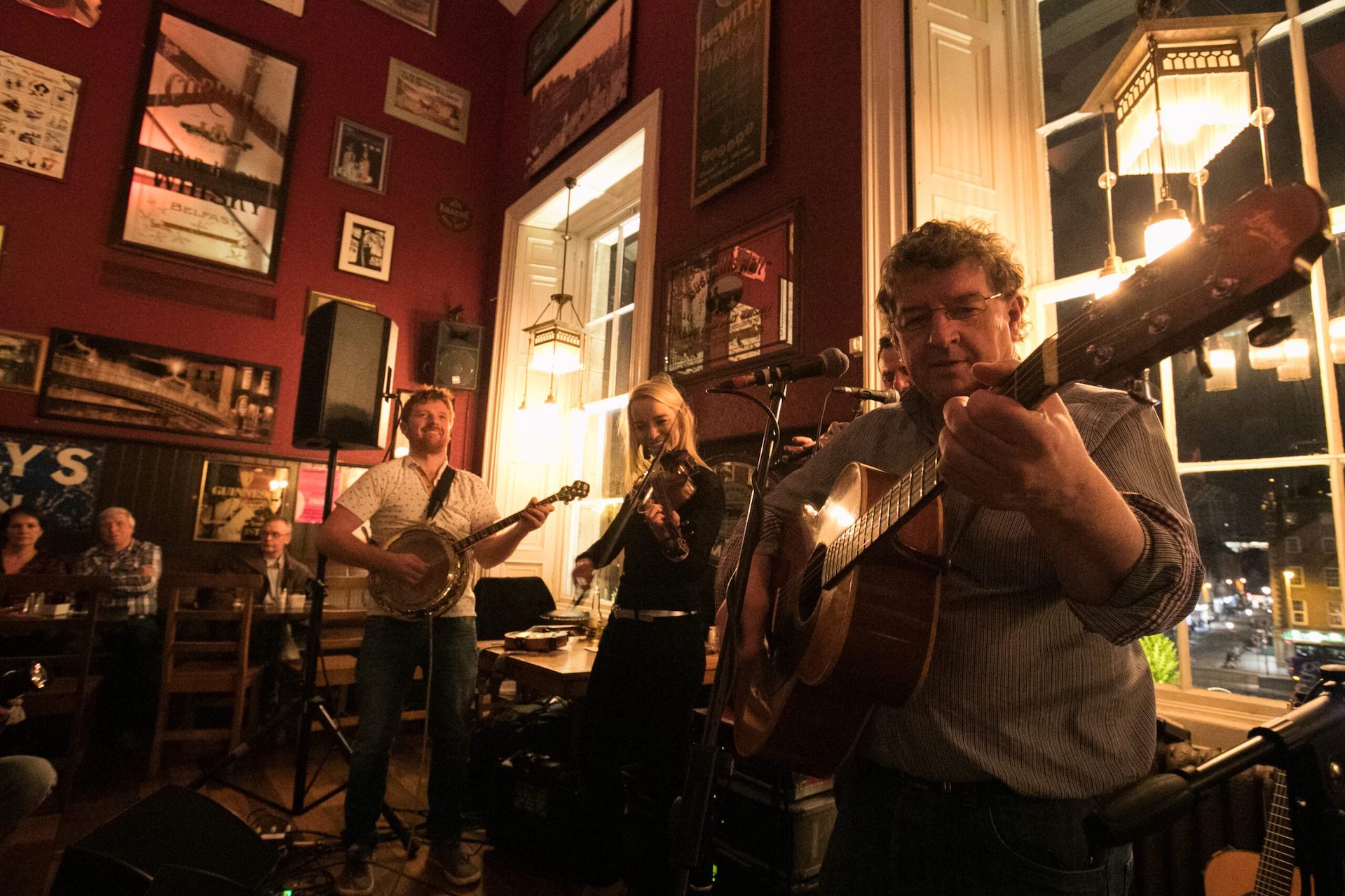 In a pub, 3 musicians are playing, standing in front of a table under 2 large windows