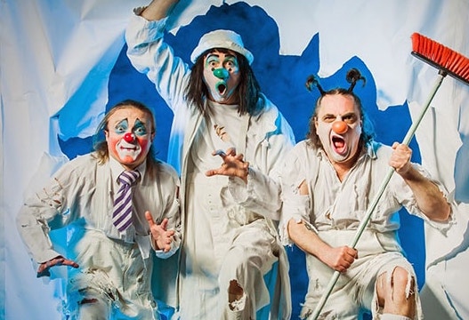 3 men in ripped white clothes with clown style faces bursting through a torn hole in a white wall, all making silly faces.