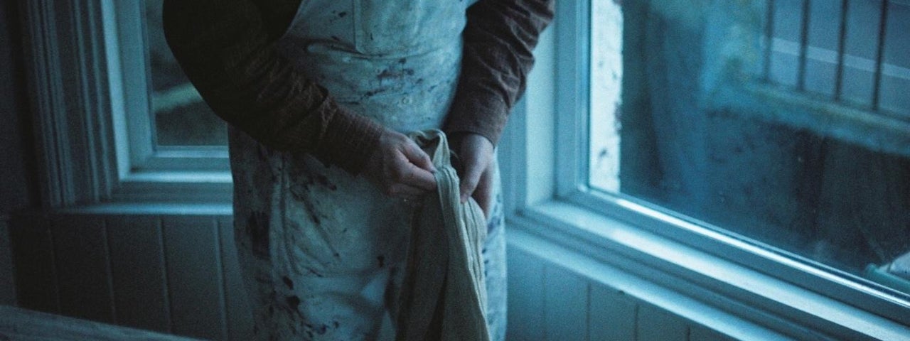 Person in old apron holding a item made of cloth, standing by a window, their head not visible.