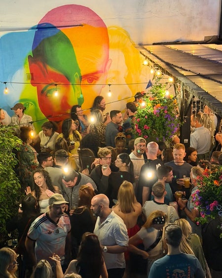 Crowds gathered in the outdoor space of a bar beneath string lights and a colourful mural backdrop