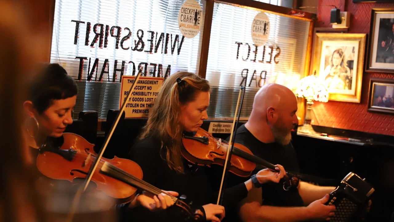 Three musicians sitting in a pub playing instruments with a window behind them