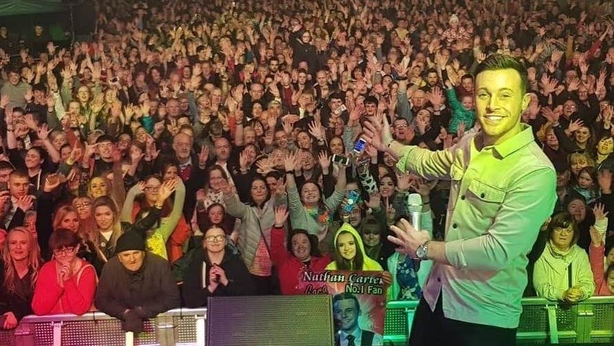 A view from the back of a stage looking out over a large crowd at night with a man to one side smiling, pointing at the audience.