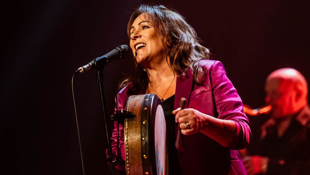 A woman singing into a mic on a stand holding a small round drum with blurred musician in the dark background.