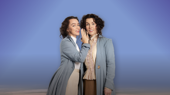 2 women in older style dress with long, grey jackets smiling at the camera, the woman on the right appearing to whisper into the ear of the other woman, against a plain blue background.