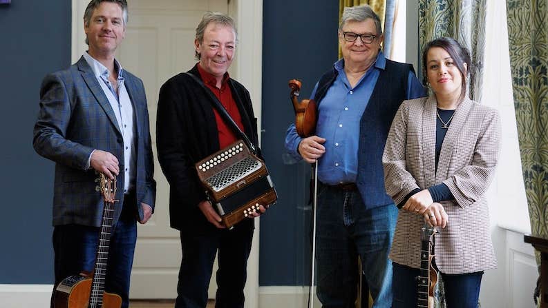4 smiling musicians standing with instruments beside a window with curtains