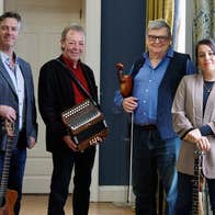 4 smiling musicians standing with instruments beside a window with curtains