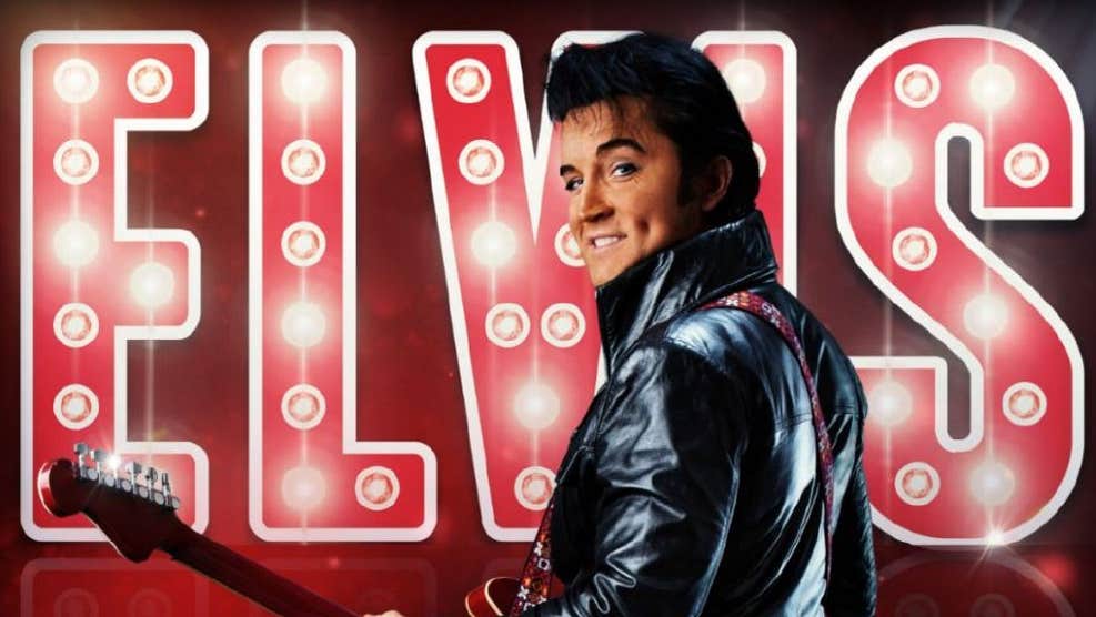 A man in leather jacket with a guitar is looking back over his left shoulder with large illuminated name behind.