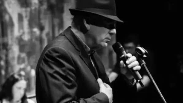 Black and white photo of man in dark suit and hat, viewed from the side, holding a mic on a stand, eyes in shadow from brim of hat.
