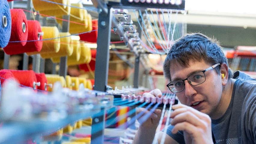 A man checking a loom with multi coloured yarn on spools