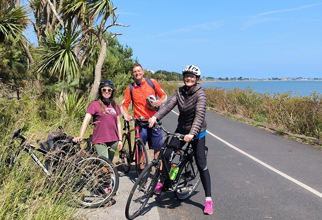 Three people with bikes on a path by the sea with trees in the background