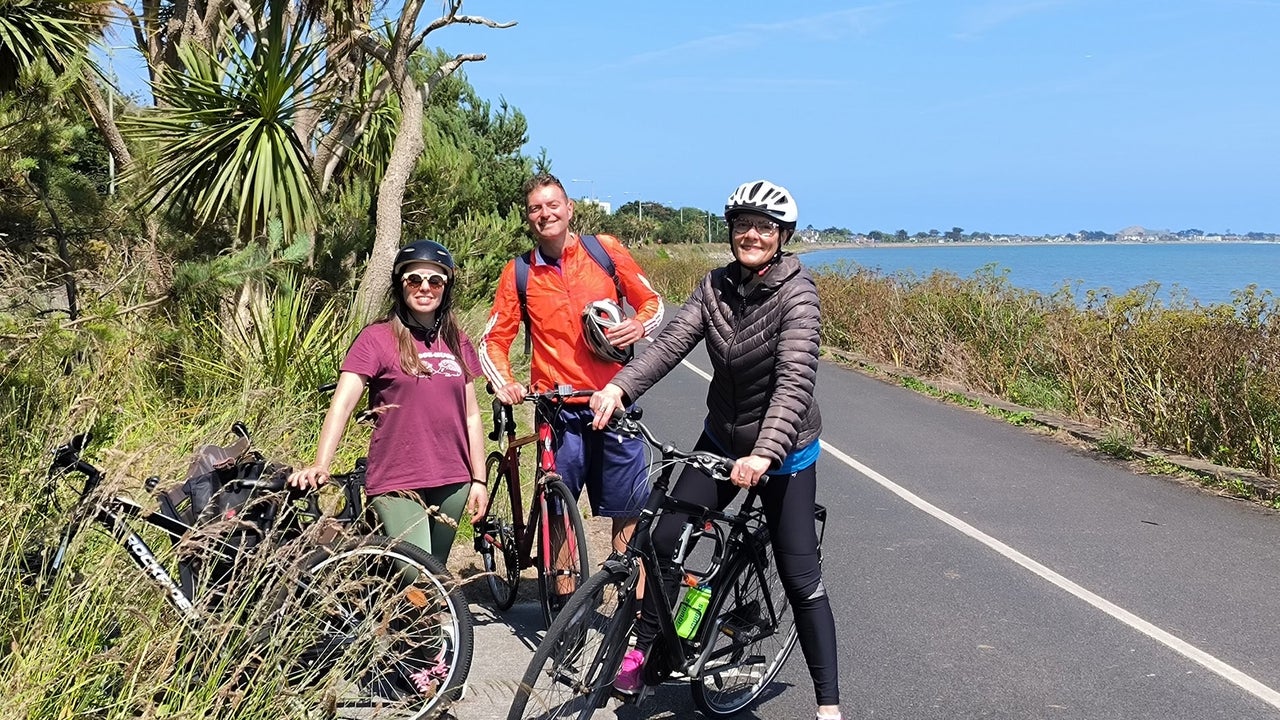 Three people with bikes on a path by the sea with trees in the background