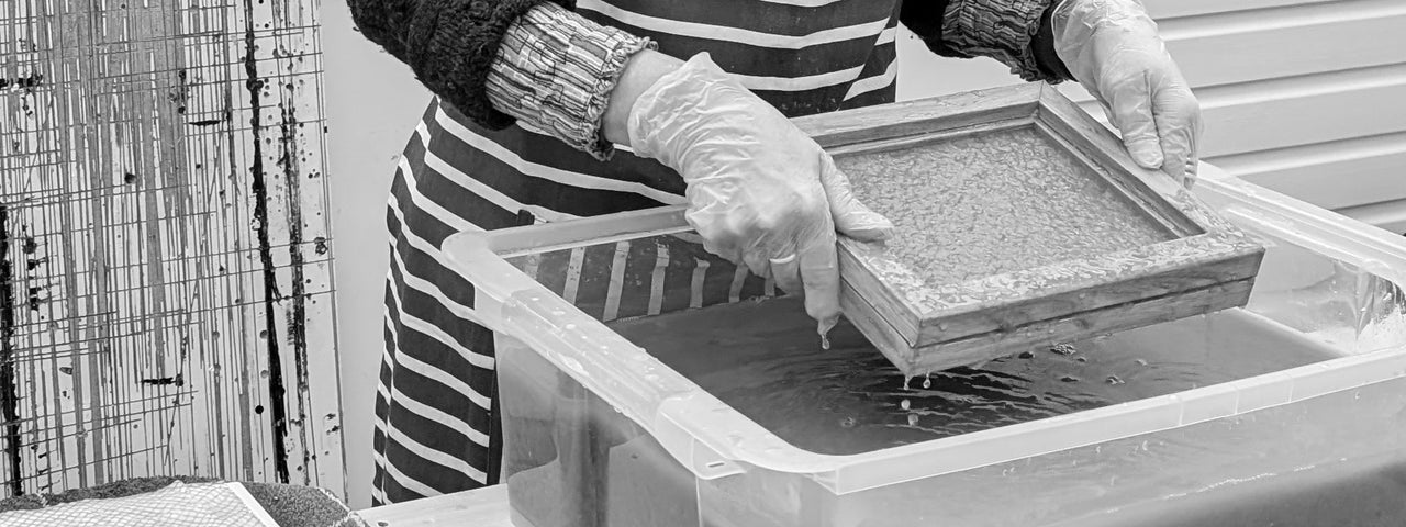 A pair of hands lifting a small wooden square frame over a large plastic container holding a dark liquid.