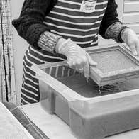A pair of hands lifting a small wooden square frame over a large plastic container holding a dark liquid.