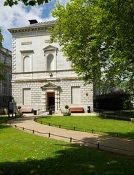 Exterior of a grey building built in a classical style with a green lawn and pathway in front