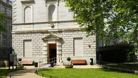 Exterior of a grey building built in a classical style with a green lawn and pathway in front
