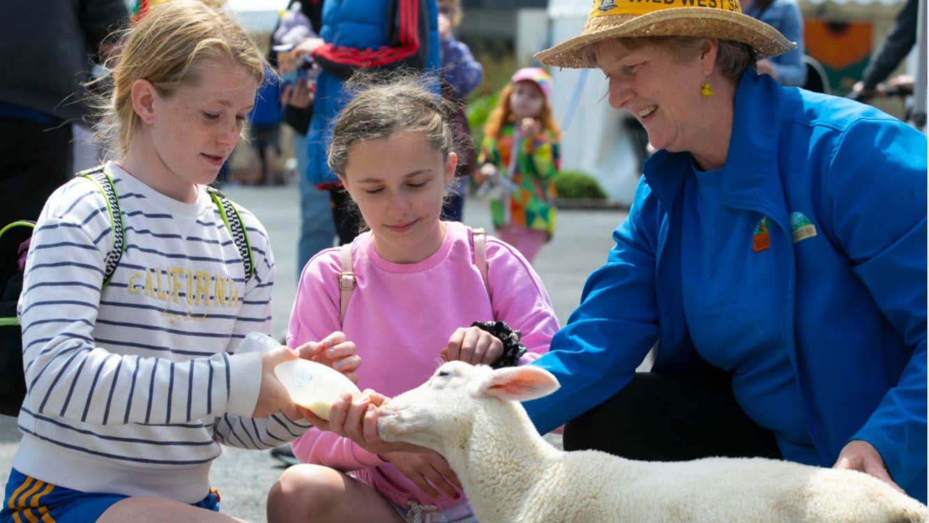 Three people crouched down with one holding a bottle feeding a lamb